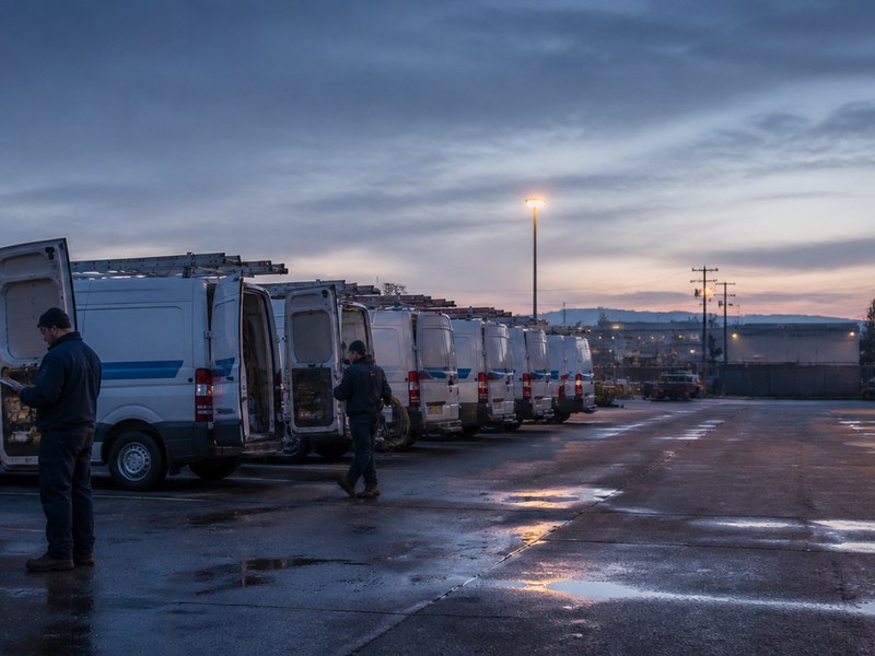 A row of service vans at a depot at dusk, two workers prepping for the day's dispatches.